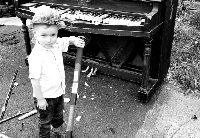 A three year old boy stands with a sledge hammer in front of a smashed piano.