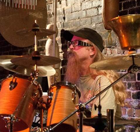 A man with long hair and bread with glasses and a hat on plays a drum set in front of a brick wall.