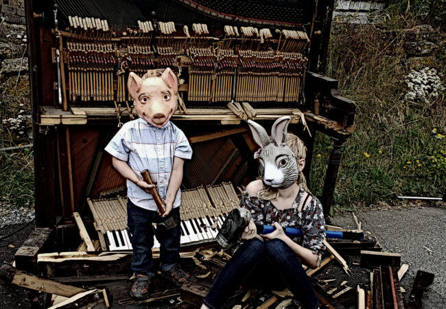 Two kids in a pig and bunny mask stand in front of a smashed piano with sledge hammers.