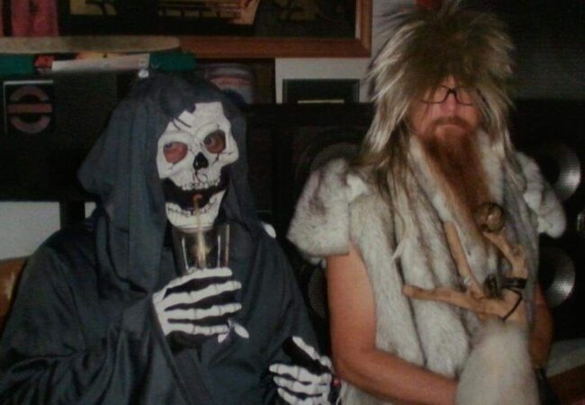 A Halloween costumed skeleton and a caveman sit and drink out of straws.