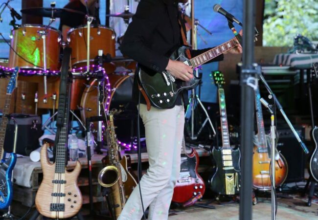 A man on stage plays a black guitar with a variety of instruments on stands behind him.