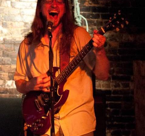 A man with long hair and glasses screams into a microphone as he plays a red guitar.