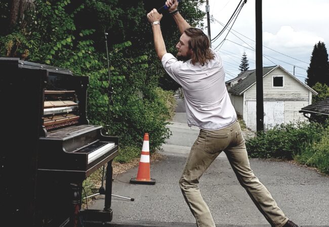 A man in an ally swings a sledge hammer above his head about to smash a piano.