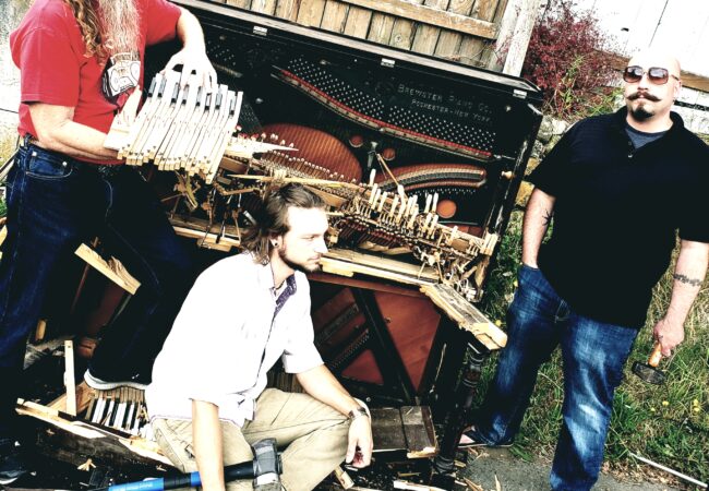 Three men standing around a smashed piano.